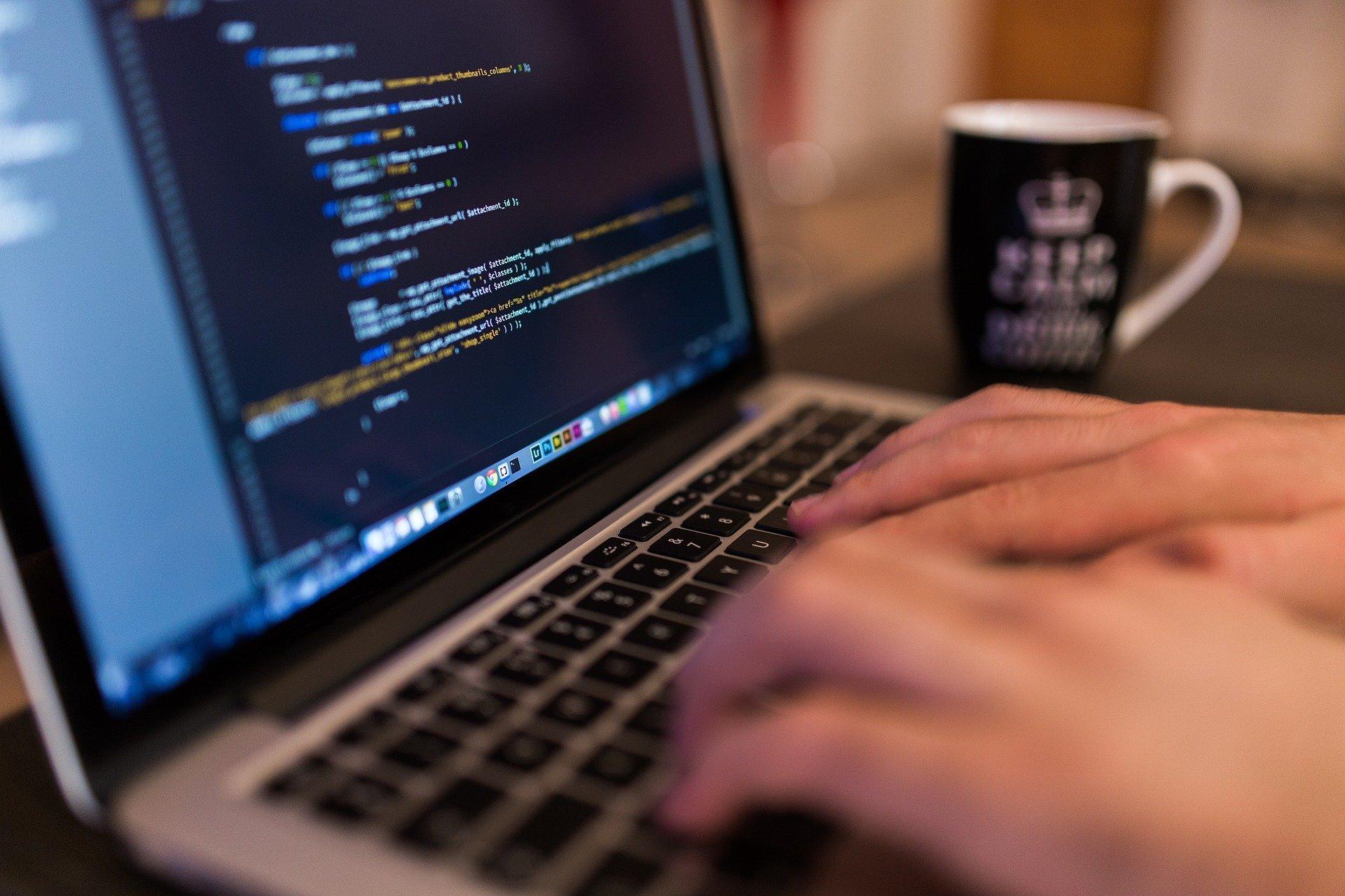 Close-up view of hands on a laptop keyword, the screen displaying colored computer code over a black background.